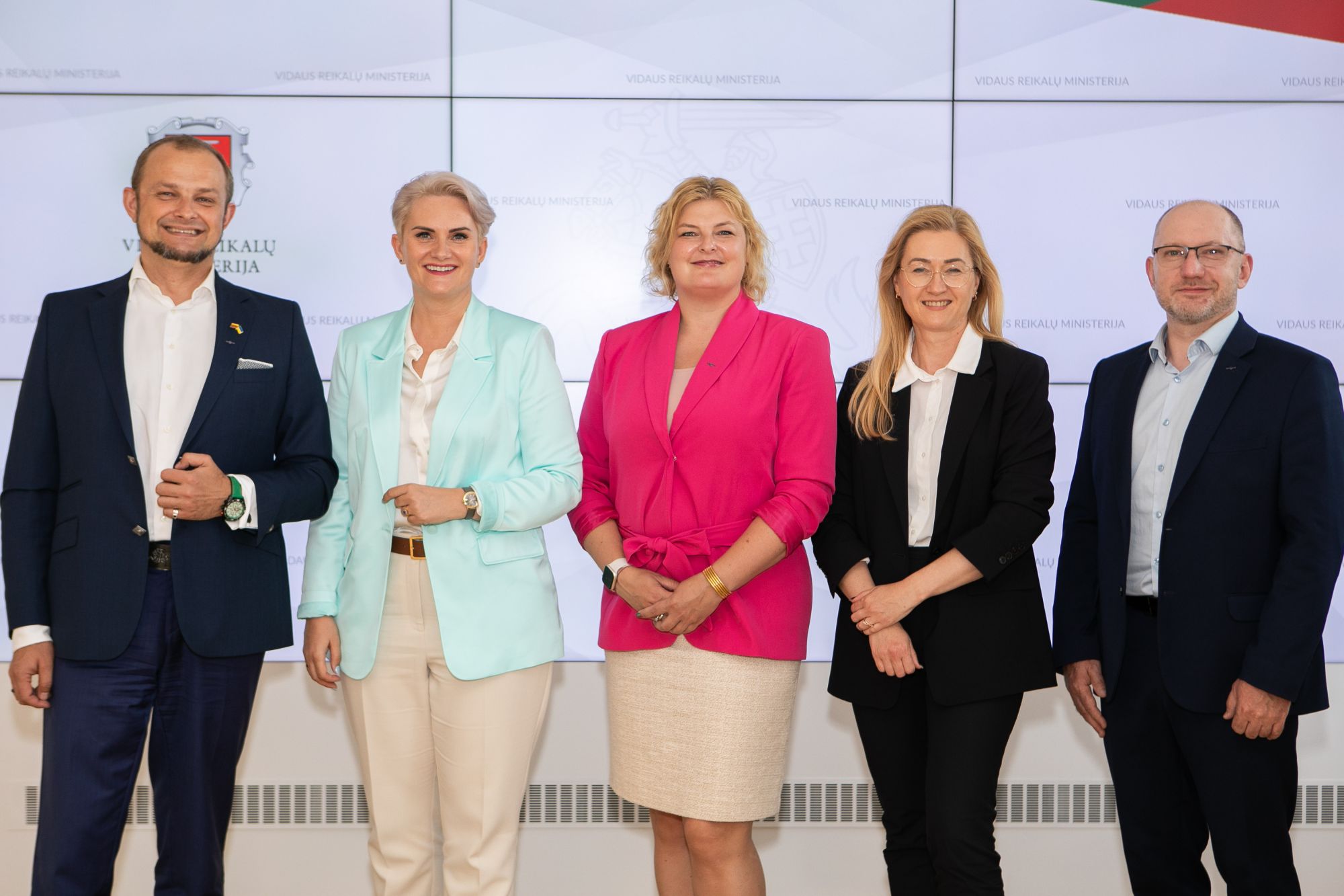 Penki asmenys stovi greta prieš foną su tekstais ir logotipa