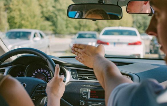 Automobilio salonas su dviem žmonėmis priekyje – vairuotojas laiko vairą, egzaminuotojas rodo ar gestikuliuoja link priekinio stiklo; pro langą matyti keli automobiliai, galimai stovintys aikštelėje ar eisme.