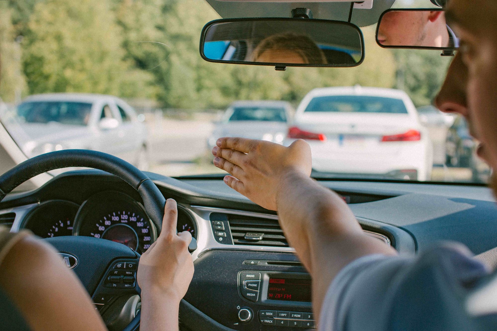 Automobilio salonas su dviem žmonėmis priekyje – vairuotojas laiko vairą, egzaminuotojas rodo ar gestikuliuoja link priekinio stiklo; pro langą matyti keli automobiliai, galimai stovintys aikštelėje ar eisme.