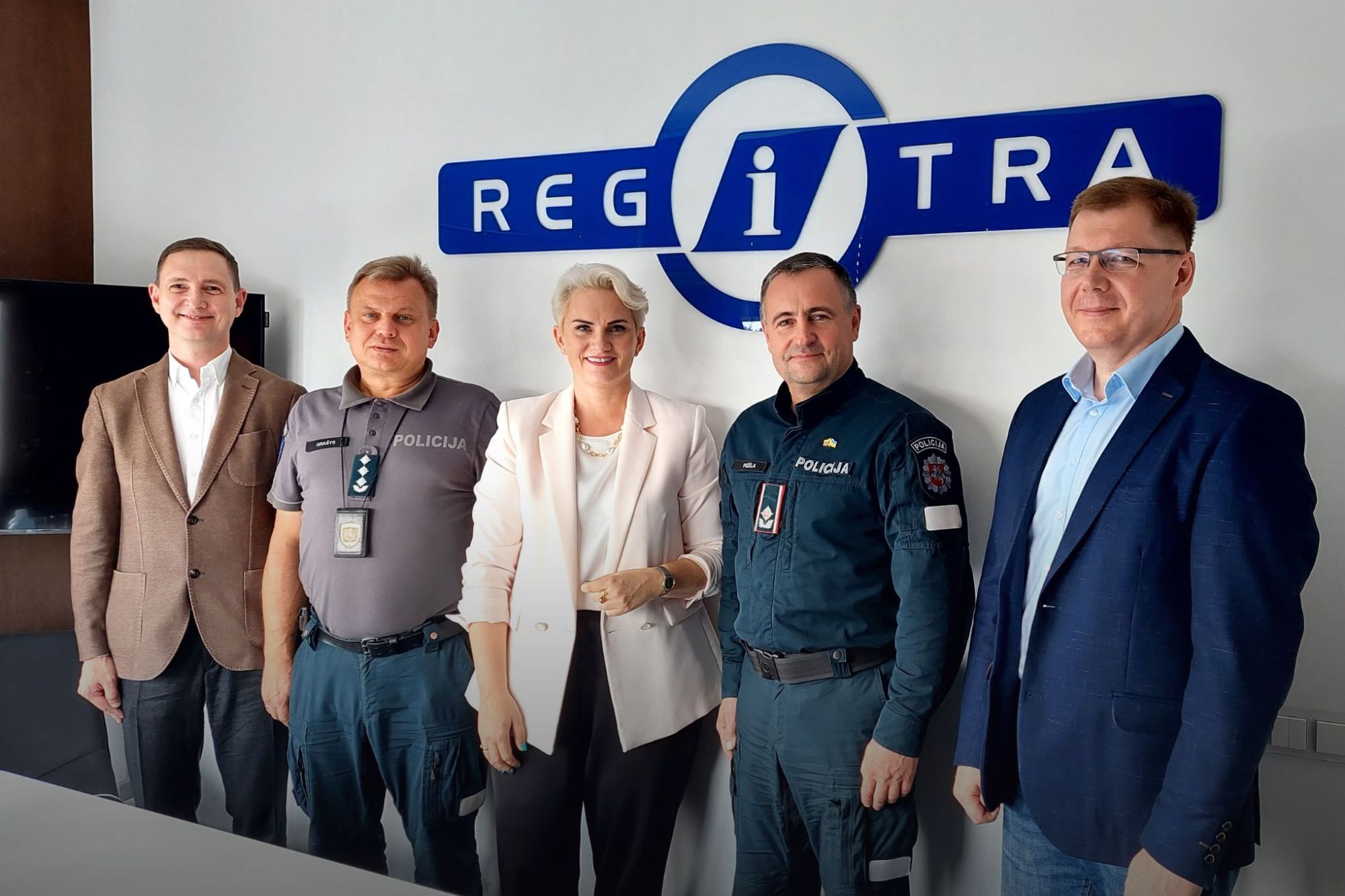 Penki asmenys stovi priešais užrašą „REGITRA“ – trys vilki policijos uniformas su užrašu „POLICIJA“, du apsirengę verslo apranga.