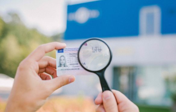 Asmuo laiko vairuotojo pažymėjimą ir padidinimo stiklą, kuriuo tikrina kortelės duomenis; fone matyti neryškus mėlynos ir baltos spalvos pastatas lauke.