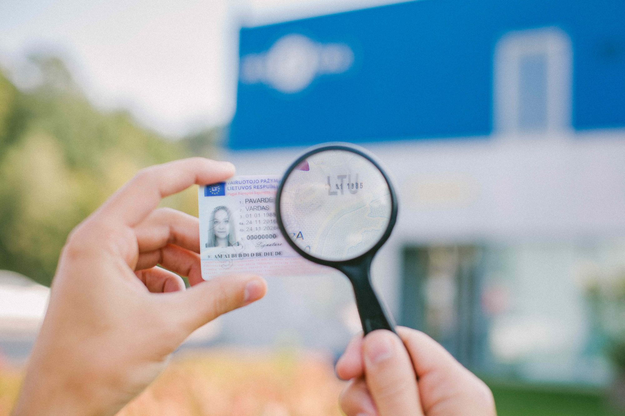 Asmuo laiko vairuotojo pažymėjimą ir padidinimo stiklą, kuriuo tikrina kortelės duomenis; fone matyti neryškus mėlynos ir baltos spalvos pastatas lauke.