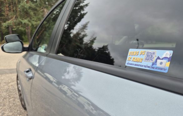 Automobilio langas su lipduku, kuriame matomas QR kodas ir tekstas „VIENS DU IK RAMU“ bei „UŽSISAKYK VAIRUOTOJO PAŽYMĖJIMĄ INTERNETU!“
