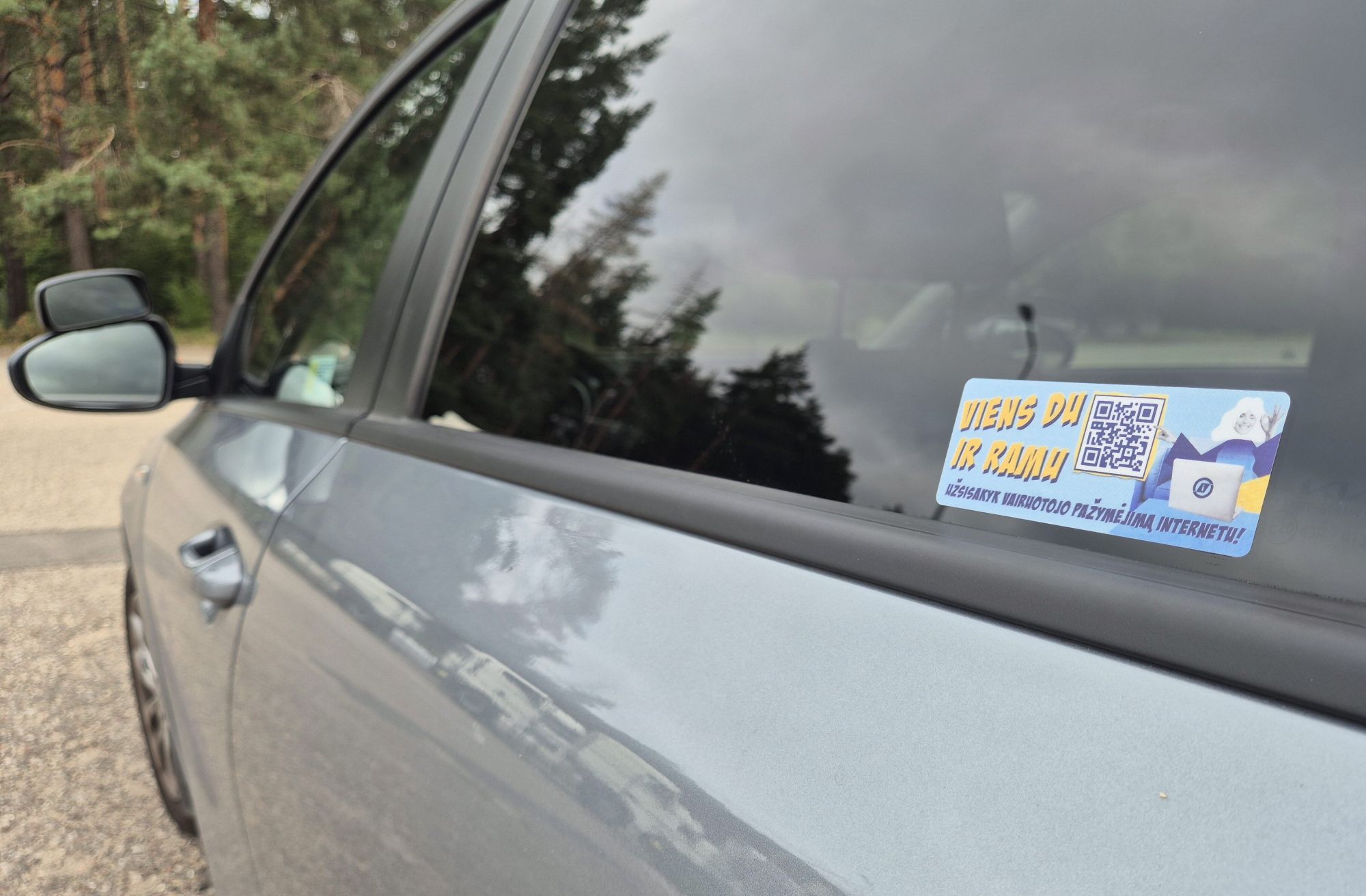 Automobilio langas su lipduku, kuriame matomas QR kodas ir tekstas „VIENS DU IK RAMU“ bei „UŽSISAKYK VAIRUOTOJO PAŽYMĖJIMĄ INTERNETU!“