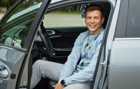 Asmuo sėdi vairuotojo vietoje atidarytomis automobilio durelėmis, vilki šviesiai mėlynus marškinėlius, pilkus kelnes ir dėvi darbo pažymėjimą ant kaklo; veidas užbluruotas.