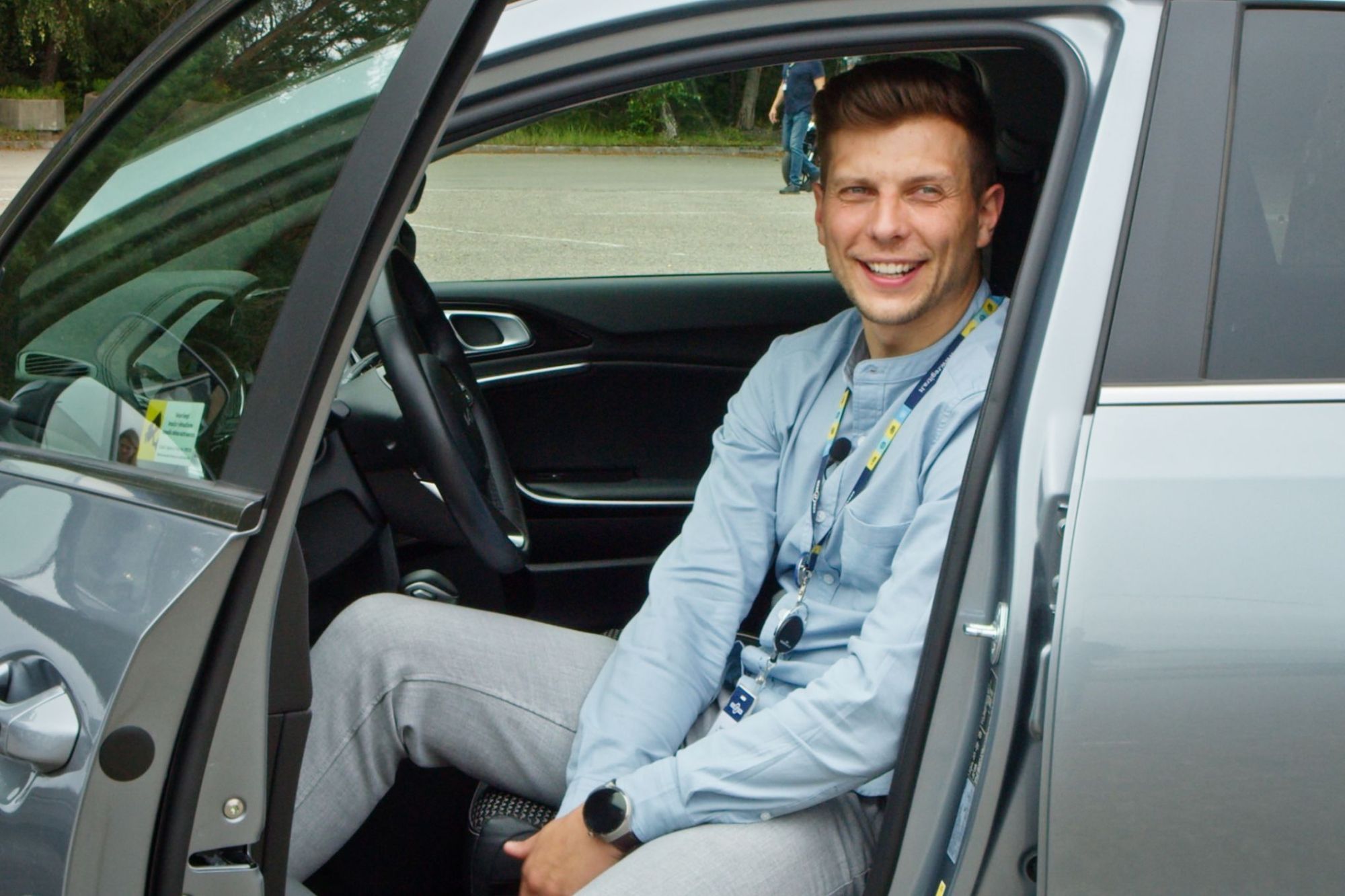 Asmuo sėdi vairuotojo vietoje atidarytomis automobilio durelėmis, vilki šviesiai mėlynus marškinėlius, pilkus kelnes ir dėvi darbo pažymėjimą ant kaklo; veidas užbluruotas.