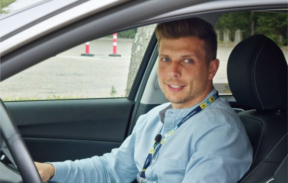 Asmuo sėdi vairuotojo vietoje atidarytomis automobilio durelėmis, vilki šviesiai mėlynus marškinėlius, pilkus kelnes ir dėvi darbo pažymėjimą ant kaklo; veidas užbluruotas.