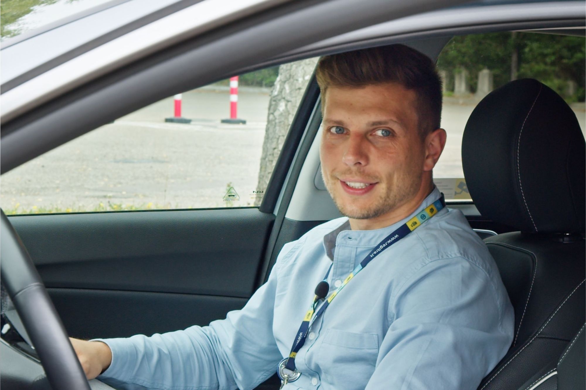 Asmuo sėdi vairuotojo vietoje atidarytomis automobilio durelėmis, vilki šviesiai mėlynus marškinėlius, pilkus kelnes ir dėvi darbo pažymėjimą ant kaklo; veidas užbluruotas.