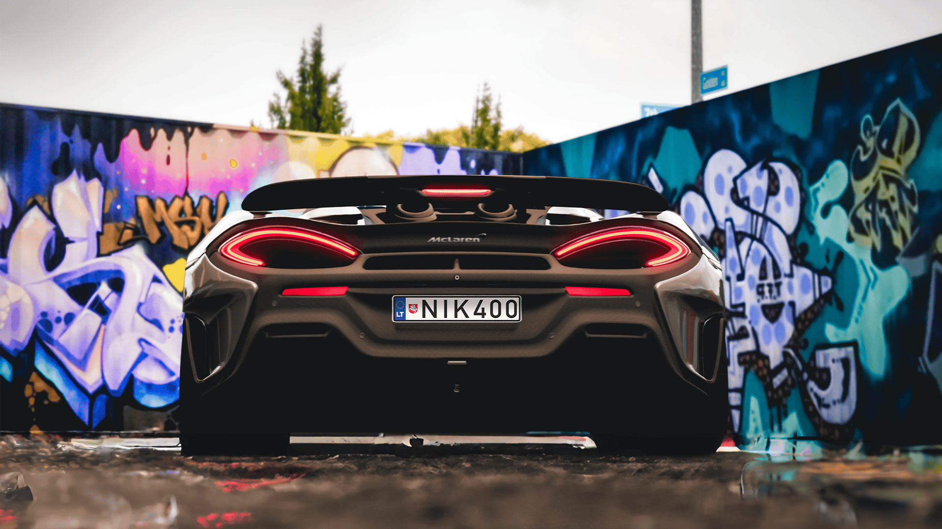 McLaren sportinis automobilis su numeriais „NIK400“, stovintis prieš spalvingą grafičiais išmargintą sieną, kurioje dominuoja mėlynos, violetinės ir geltonos spalvos.