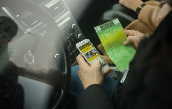 Asmuo sėdi automobilyje, rankose laiko išmanųjį telefoną ir žalią dokumentą; telefono ekrane matoma programėlė su dviejų žmonių atvaizdu. Fone – automobilio prietaisų skydelis, vairas ir keleivis su rusvu paltu.