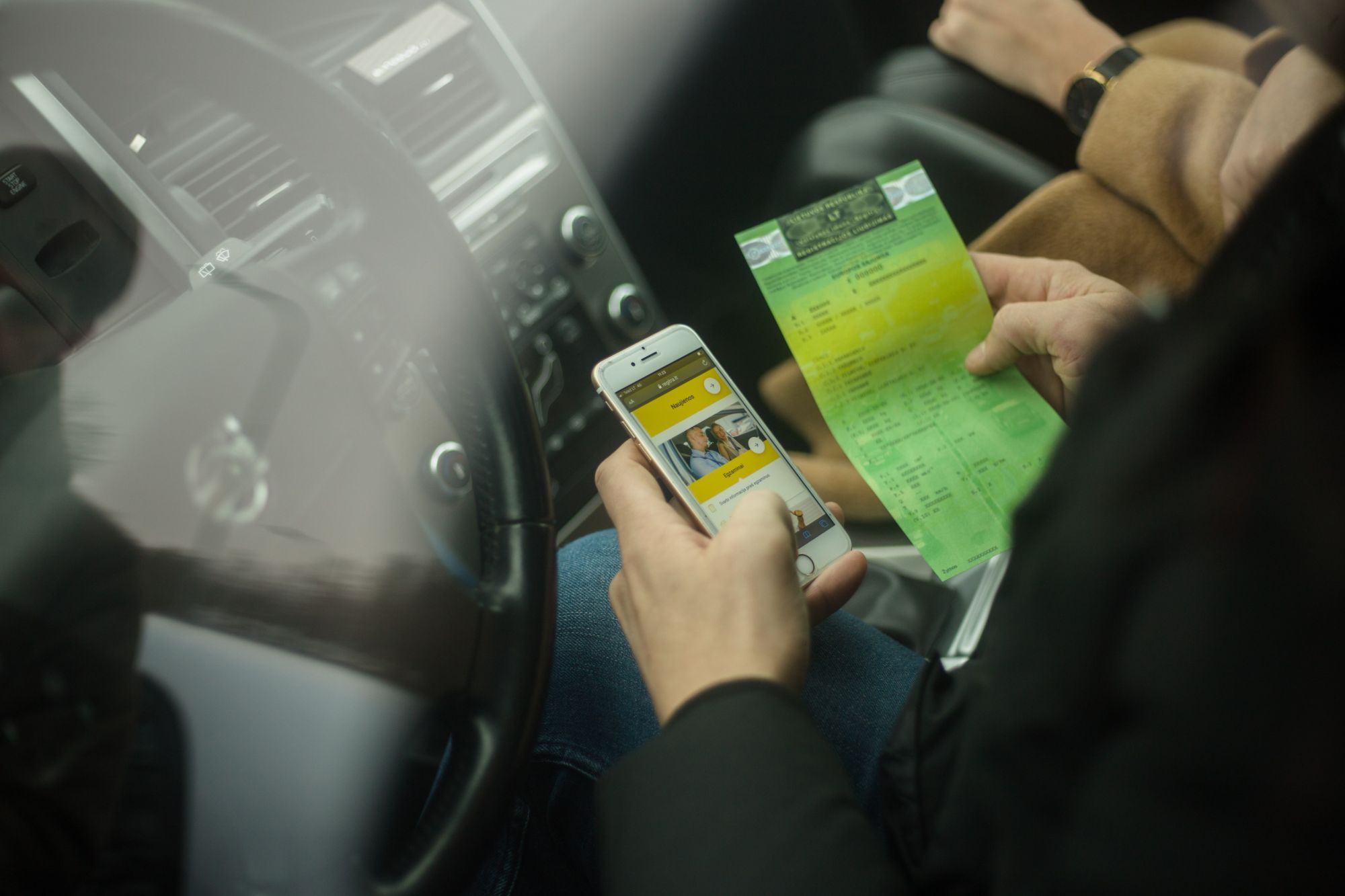 Asmuo sėdi automobilyje, rankose laiko išmanųjį telefoną ir žalią dokumentą; telefono ekrane matoma programėlė su dviejų žmonių atvaizdu. Fone – automobilio prietaisų skydelis, vairas ir keleivis su rusvu paltu.