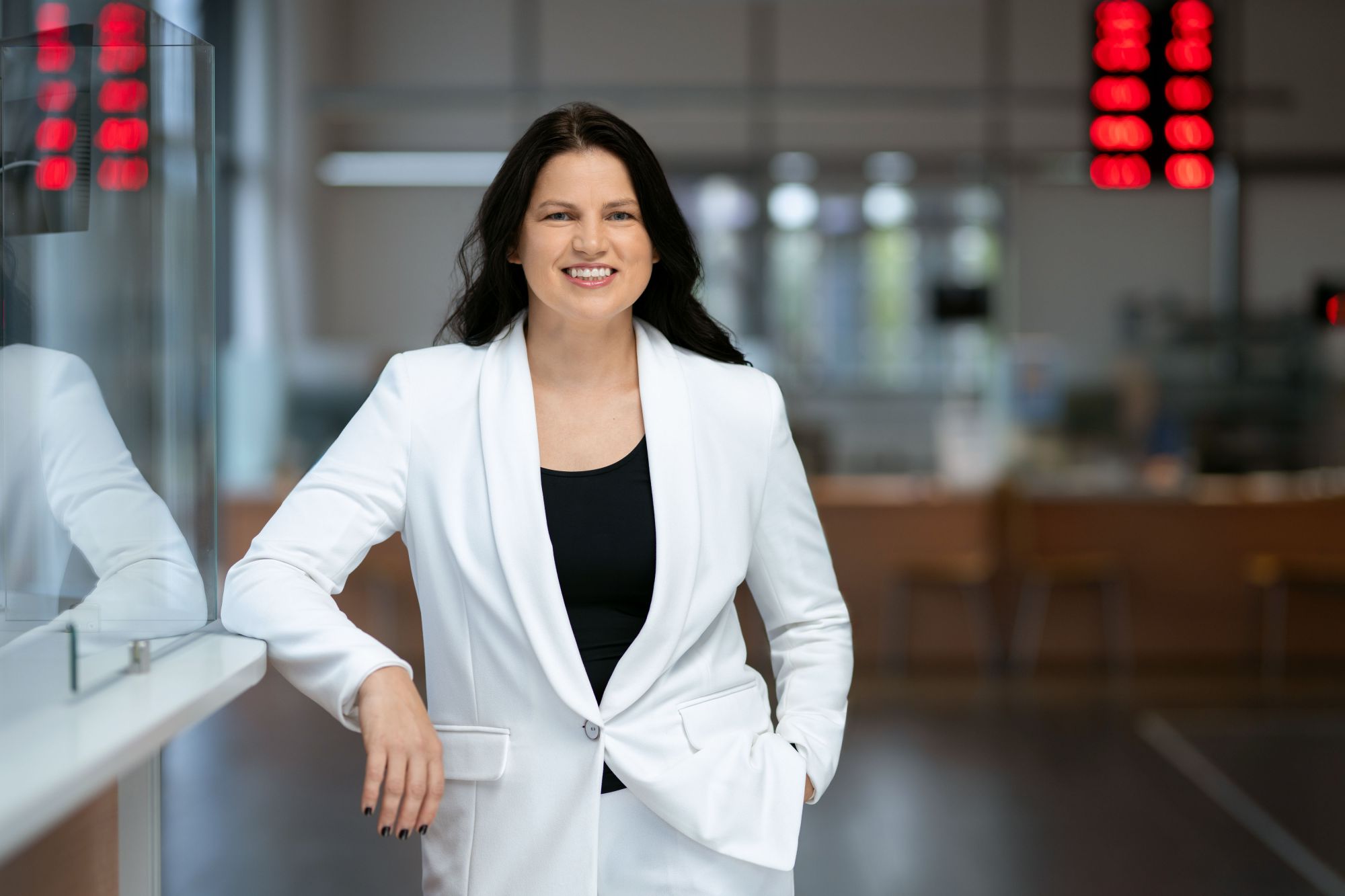 Asmuo su baltu švarku ir juodu viršumi stovi patalpoje, viena ranka atsirėmusi, kita kišenėje; fone matyti neryškūs baldai ir skaitmeniniai ekranai su raudonais skaičiais.