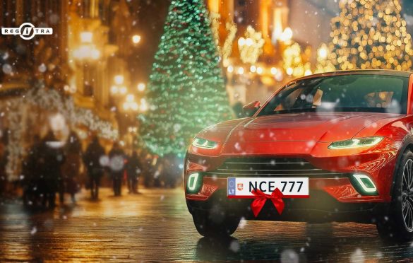 Šventiškai papuošta gatvė su apšvišta Kalėdų eglute fone; priekyje – raudonas automobilis su įjungtomis šviesomis ir lietuviškais numeriais „NCE 777“.