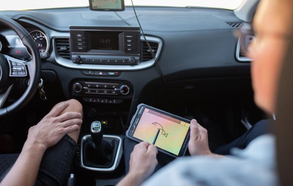 Automobilio salonas, kuriame du žmonės – vienas laiko planšetę su brėžiniu ar schema, kito rankos matomos prie pavarų svirties; matomas vairas, multimedijos sistema ir oro kondicionavimo valdymas, ekranas rodo laiką 12:27.