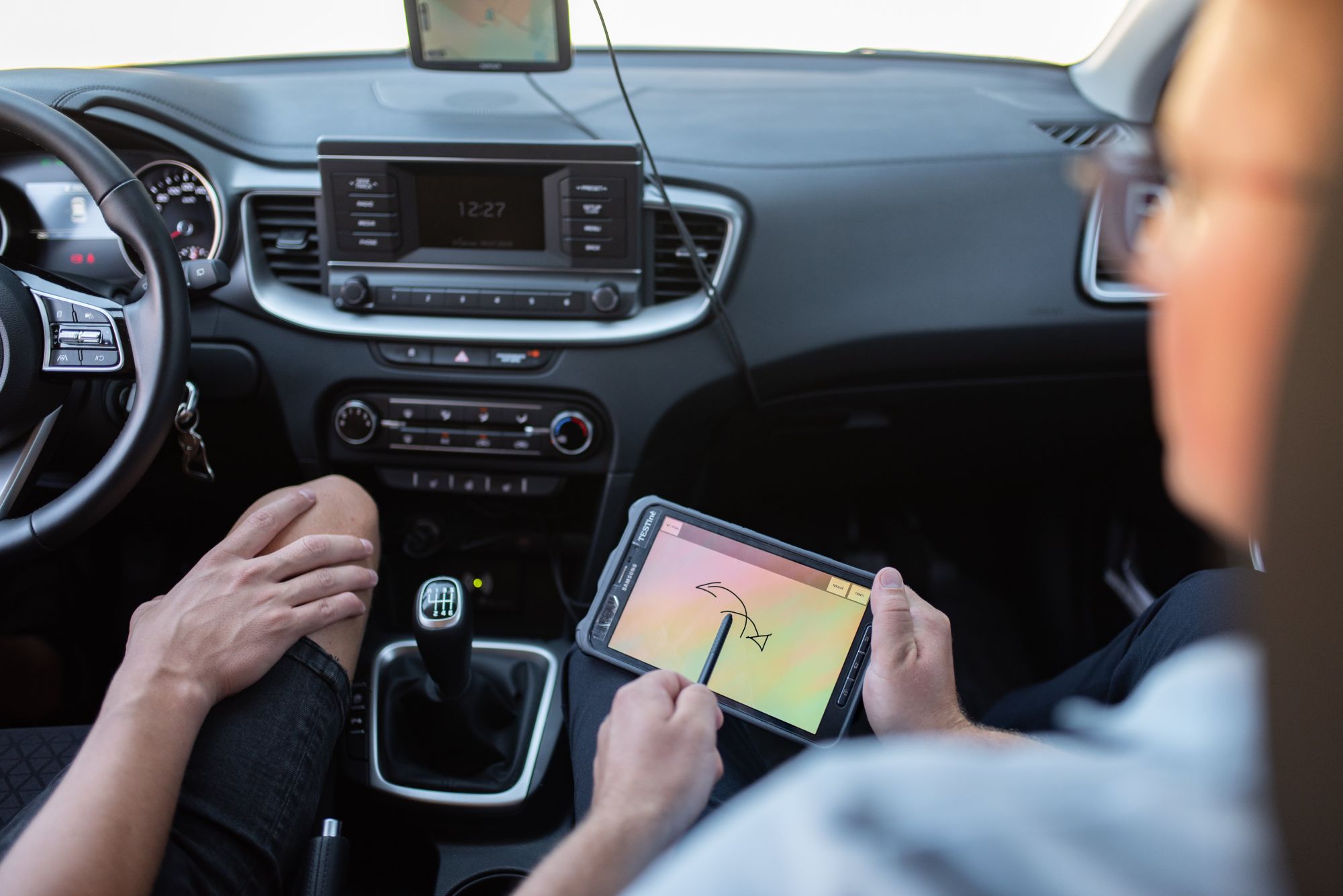 Automobilio salonas, kuriame du žmonės – vienas laiko planšetę su brėžiniu ar schema, kito rankos matomos prie pavarų svirties; matomas vairas, multimedijos sistema ir oro kondicionavimo valdymas, ekranas rodo laiką 12:27.