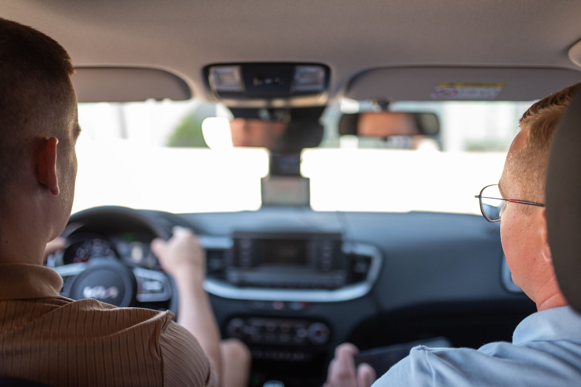 Vaizdas iš automobilio galinės sėdynės – priekyje du žmonės, vienas vairuoja, kitas šalia; matomas prietaisų skydelis, vairas ir priekinio stiklo viršuje pritvirtintas GPS įrenginys