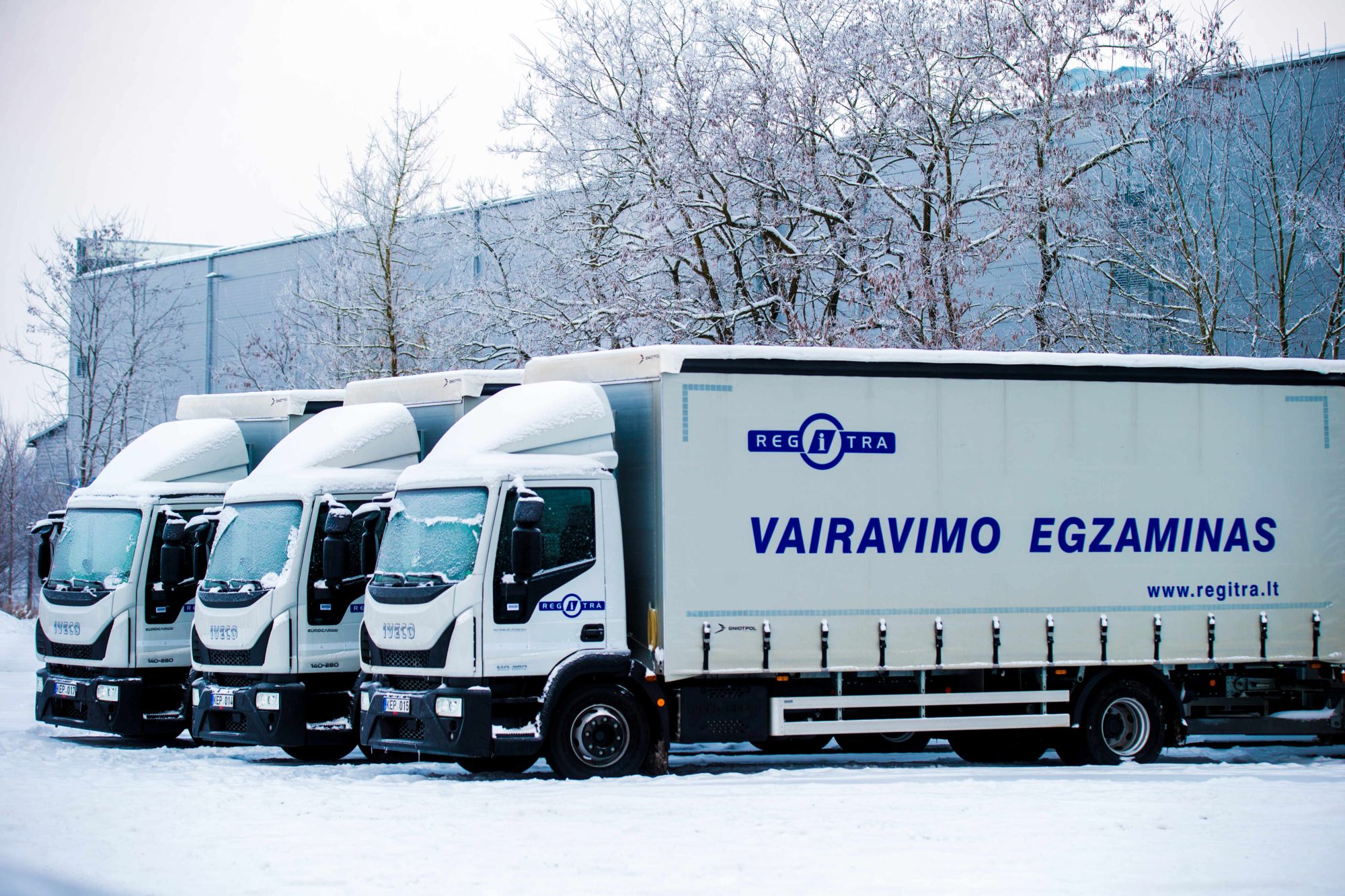 Keturi balti „Regitra“ sunkvežimiai stovi apsnigtame plote. Ant jų matomas užrašas „VAIRAVIMO EGZAMINAS“ ir svetainės adresas www.regitra.lt. Fone – apsnigti medžiai ir didelis pastatas.