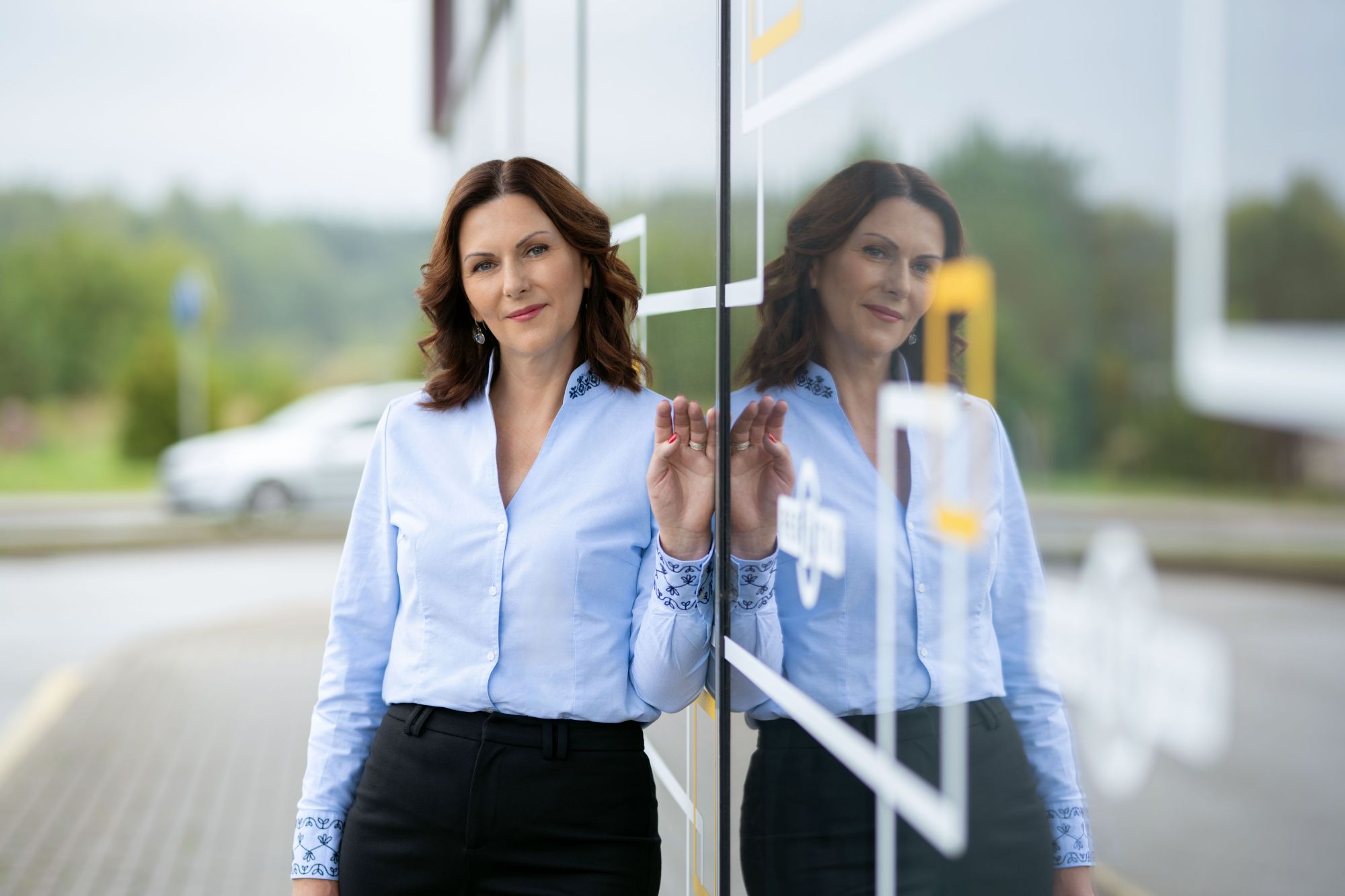 Moteris su šviesiai mėlynais marškiniais ir tamsiomis kelnėmis stovi prie atspindinčios stiklinės sienos. Veidas uždengtas kvadratine neryškumo zona, matomas atspindys stikle.