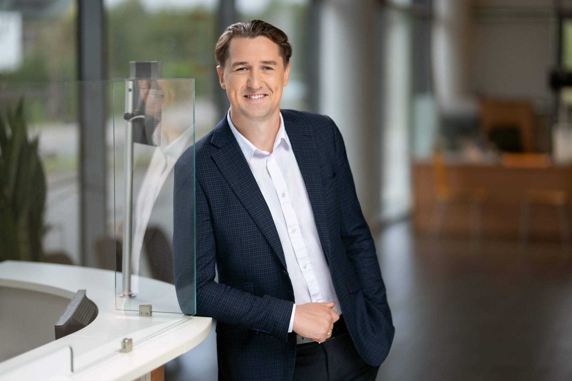 Asmuo su tamsiai mėlynu kostiumu ir baltais marškiniais atsirėmęs į stiklinę pertvarą biuro aplinkoje. Fone matomi dideli langai ir neryškūs biuro baldai. Veidas neatskleistas.