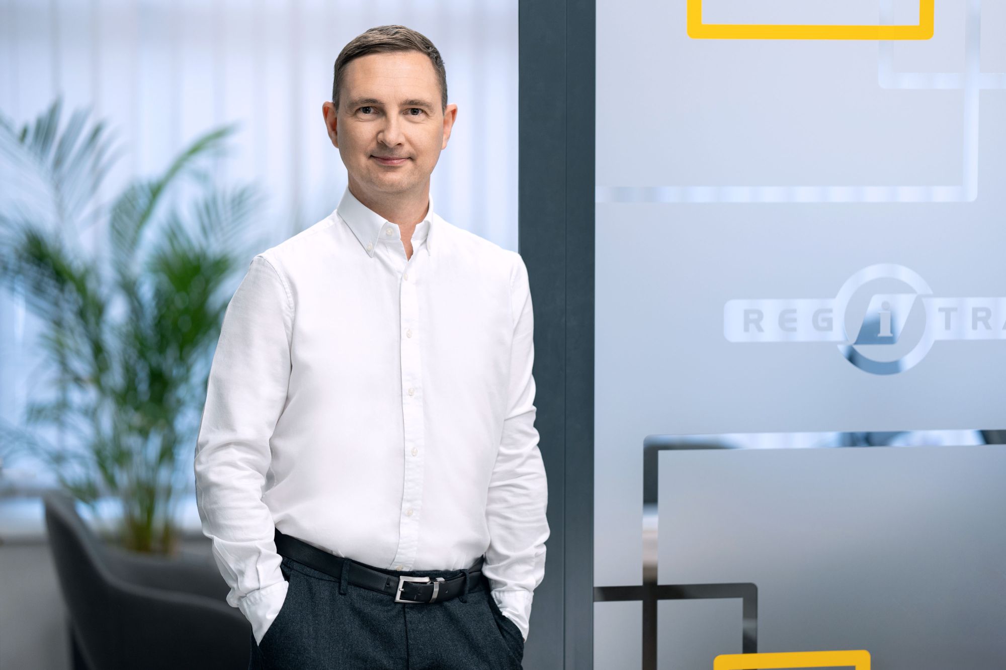 Asmuo su baltais marškiniais ir tamsiomis kelnėmis stovi patalpoje, rankos kišenėse. Fone matomi augalai ir matinio stiklo pertvaros su dalinai matomu užrašu „REGI“. Veidas neryškus.