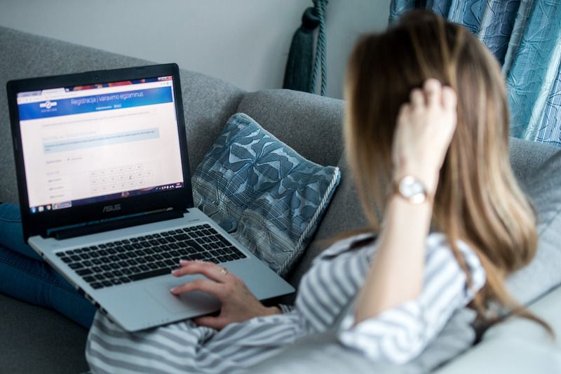 Asmuo su dryžuotais marškiniais sėdi ant sofos ir naudojasi ASUS nešiojamuoju kompiuteriu, ekrane matoma svetainė „Registracija į vairavimo egzaminus“.