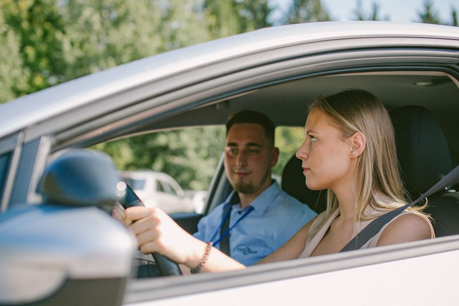 Du žmonės sėdi automobilyje, vairuotojas laiko vairą, keleivis sėdi šalia