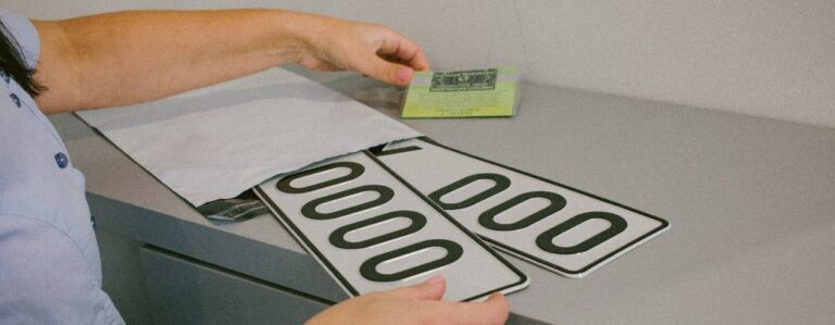 Šioje nuotraukoje matomas žmogus, tvarkantis transporto priemonių numerius su skaičiais „0000“. Asmuo taip pat laiko žalią kortelę arba dokumentą, o ant stalo yra vokas arba siuntinys, iš kurio numeriai išimami.