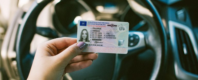 Rankoje laikomas lietuviškas vairuotojo pažymėjimas, fone matomas automobilio vairas
