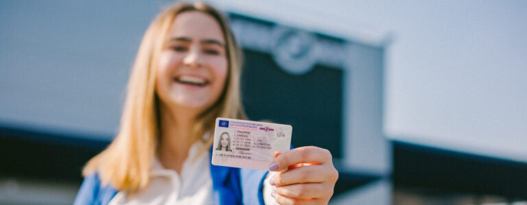 Šioje nuotraukoje matyti žmogus, stovintis priešais pastatą ir laikantis vairuotojo pažymėjimą.