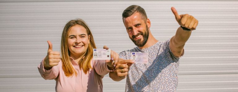 Du žmonės, vyras ir moteris, rodo vairuotojo pažymėjimus ir kelia nykščius prie šviesios sienos