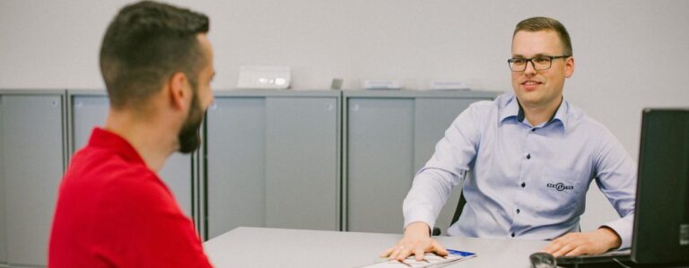 Šioje nuotraukoje matomi du žmonės, sėdintys vienas priešais kitą prie stalo, greičiausiai biuro aplinkoje. Kairėje – asmuo su raudonais marškinėliais, dešinėje – su šviesiai mėlynais marškiniais, ant kurių matomas Regitros logotipas.