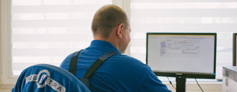 Šioje nuotraukoje matomas žmogus, sėdintis prie stalo ir žiūrintis į kompiuterio monitorių. Jis vilki mėlynus marškinėlius, ant kurių nugaros matomas užrašas „REGITRA“