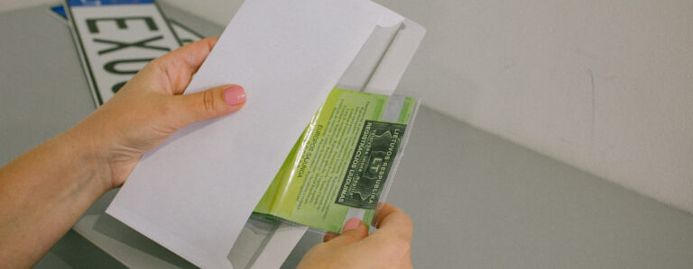 Šioje nuotraukoje matomos rankos, laikančios voką ir įdedančios arba išimančios žalią kortelę su tekstu. Fone dalinai matomas automobilio numeris, kuriame įskaitomos raidės „EXU“.