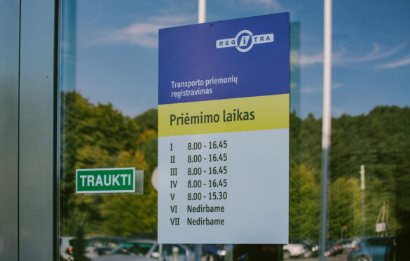 Šioje nuotraukoje matomas informacinis stendas ant stiklinės durų su „REGITRA“ logotipu viršuje. Po juo pateikta informacija apie transporto priemonių registravimo darbo laiką: I–IV: 8.00 – 16.45 V: 8.00 – 15.30 VI–VII: nedirbama