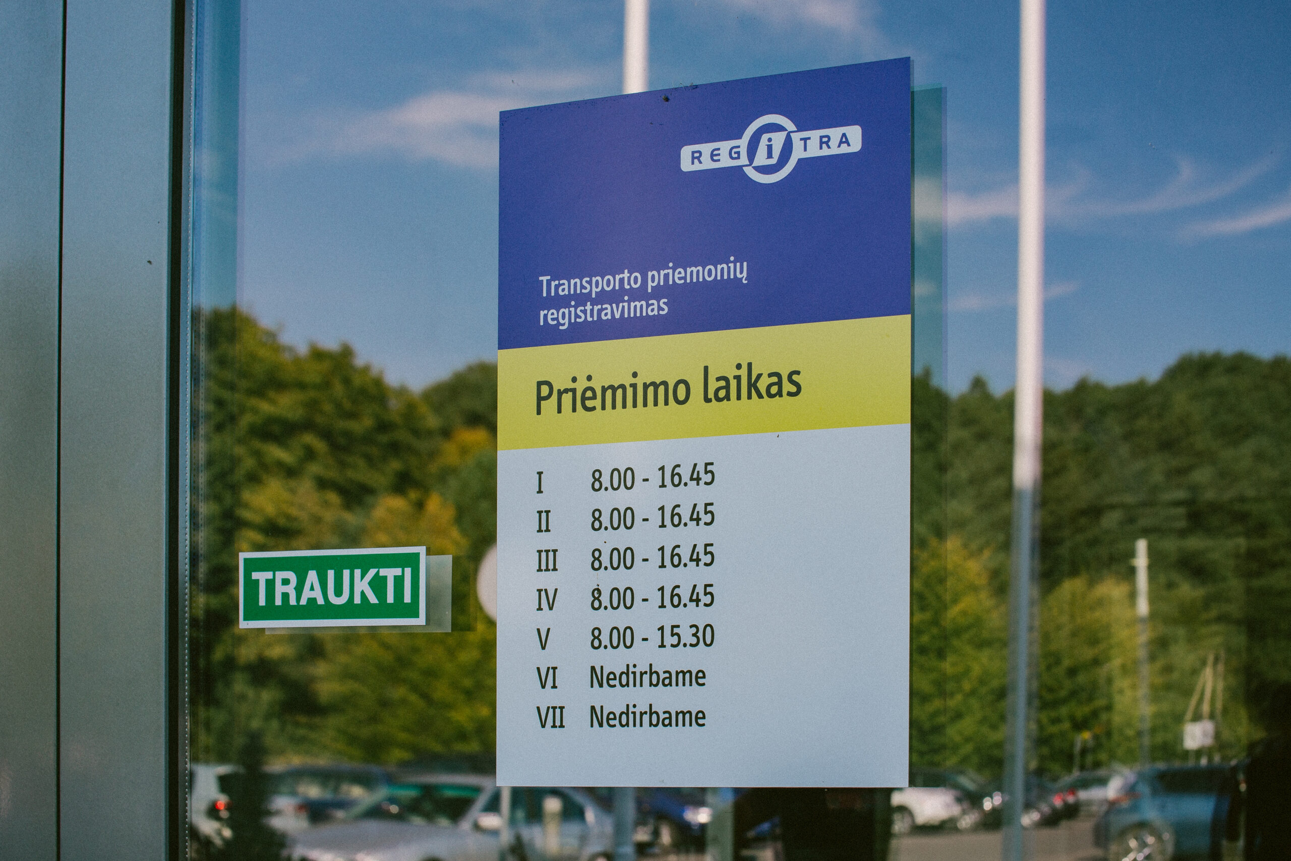 Šioje nuotraukoje matomas informacinis stendas ant stiklinės durų su „REGITRA“ logotipu viršuje. Po juo pateikta informacija apie transporto priemonių registravimo darbo laiką: I–IV: 8.00 – 16.45 V: 8.00 – 15.30 VI–VII: nedirbama