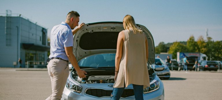 Praktikos egzamino pradžioje du žmonės stovi prie automobilio, kuriam pakeltas kapotas, ir apžiūrinėja variklį ir kitas dalis