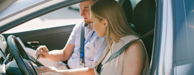 Du žmonės sėdi automobilio priekinėse sėdynėse, vienas aiškina kažką su rašikliu, kitas – prisegtas saugos diržu