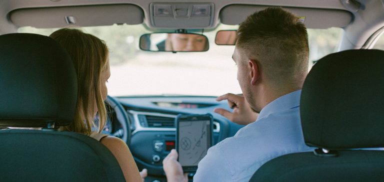 Praktikos vairavimo egzaminas – automobilyje sėdi moteris, kuri laiko egzaminą ir egzaminuotojas, egzaminuotojas rodo maršrutą navigacijoje