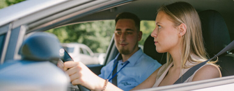 Du žmonės sėdi automobilyje, vairuotojas laiko vairą, keleivis sėdi šalia