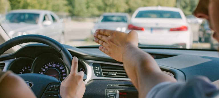 Du žmonės automobilio priekyje – vairuotojas rodo pirštu, keleivis gestikuliuoja. Pro langą matyti kiti automobiliai