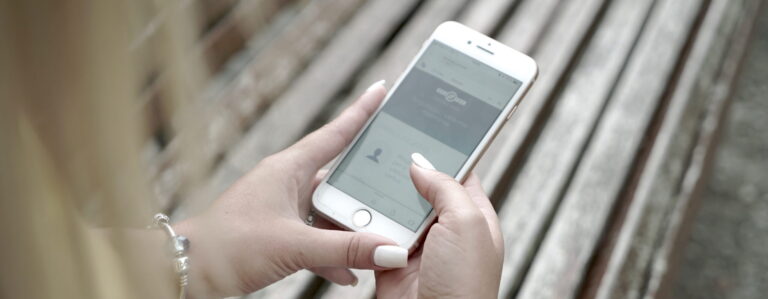 Šioje nuotraukoje matomas žmogus, sėdintis ant medinio suoliuko ir laikantis išmanųjį telefoną. Ekrane matoma Regitros puslapis.