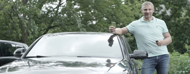 Šioje nuotraukoje matomas žmogus, stovintis šalia automobilio. Vienoje rankoje jis laiko išmanųjį telefoną, o kitoje – automobilio raktą.