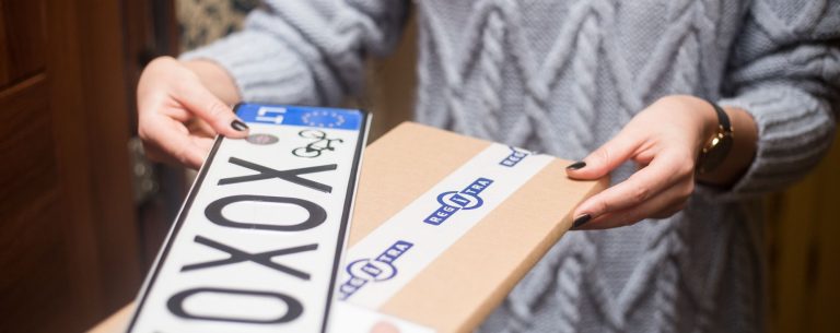 Asmuo su pilku megztiniu laiko numerio lentelę „XOXOX“ ir pakuotę su užrašu „REGITRA“.