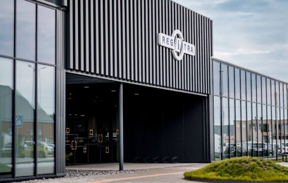 Šioje nuotraukoje matyti modernus pastatas su juodu fasadu ir dideliais stikliniais langais. Virš įėjimo yra užrašas „REGITRA“, nurodantis, kad tai greičiausiai vienas iš „Regitros“ padalinių. Langų atspindyje ir fone matyti keletas automobilių, o prie įėjimo – šiek tiek žalumos. Dangus dalinai debesuotas.