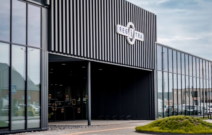 Šioje nuotraukoje matyti modernus pastatas su juodu fasadu ir dideliais stikliniais langais. Virš įėjimo yra užrašas „REGITRA“, nurodantis, kad tai greičiausiai vienas iš „Regitros“ padalinių. Langų atspindyje ir fone matyti keletas automobilių, o prie įėjimo – šiek tiek žalumos. Dangus dalinai debesuotas.