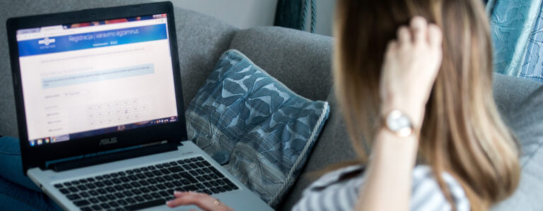 Šioje nuotraukoje matomas žmogus, sėdintis ant sofos ir naudojantis ASUS nešiojamąjį kompiuterį. Ekrane atidarytas puslapis su antrašte „Registracija į vairavimo egzaminus“