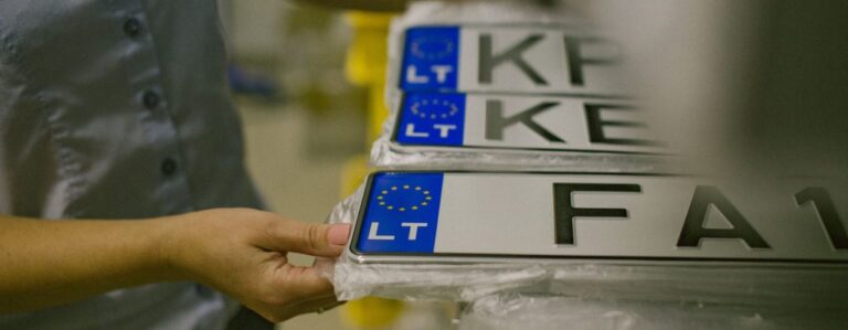 Paveikslėlyje matomas asmuo, laikantis krūvą Europos Sąjungos stiliaus automobilių numerių. Numeriuose yra mėlynas ES ženklas su geltonomis žvaigždėmis kairėje pusėje ir raidės „LT“, nurodančios Lietuvą. Matomi numeriai yra „KP“, „KE“ ir „FA1“.