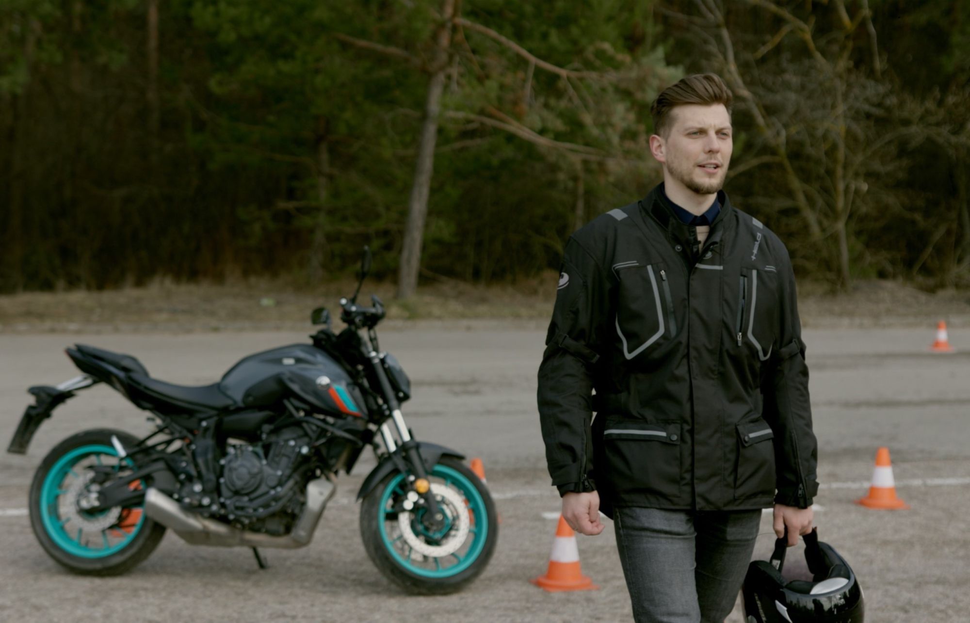 Asmuo su juodu švarku laiko šalmą lauke, fone matomas juodas motociklas su žalsvais ratlankiais ir keli oranžiniai kelio kūgiai.