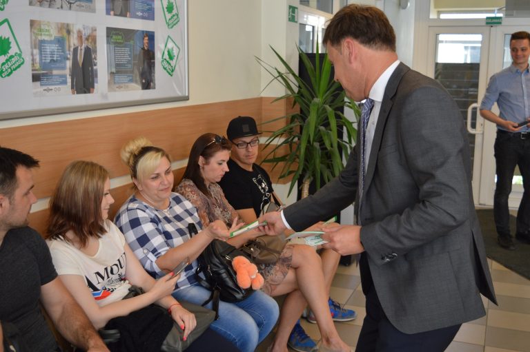 Grupė žmonių sėdi ant suoliuko laukimo zonoje, o vyras kostiumu stovi ir dalina dokumentus ar lankstinukus. Už jų – plakatai ant sienos ir didelis augalas.