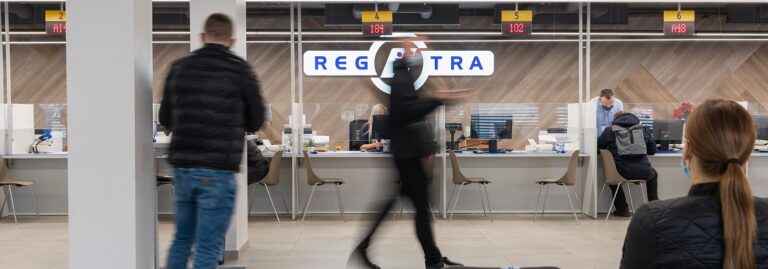 rie aptarnavimo langelių sėdi klientai ir darbuotojai. Virš langelių – skaitmeniniai ekranai su numeriais ir raidėmis, rodančiais eilės numerius. Fone matomas užrašas „REGITRA“